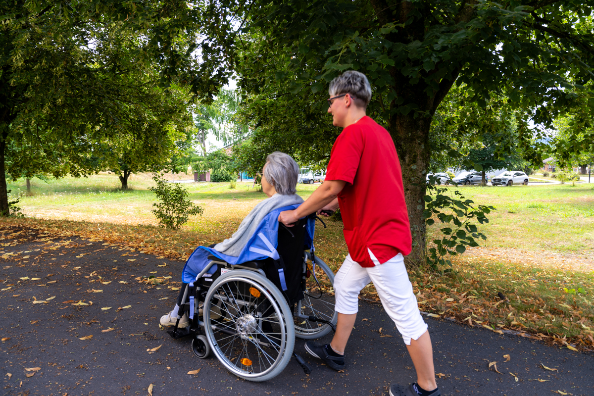 eine Pflegerin schiebt eine Bewohnerin im Rollstuhl durch den Park